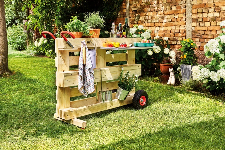 Eine mobile Gartenbar aus Holz mit Rädern und einer Arbeitsfläche steht im Garten.