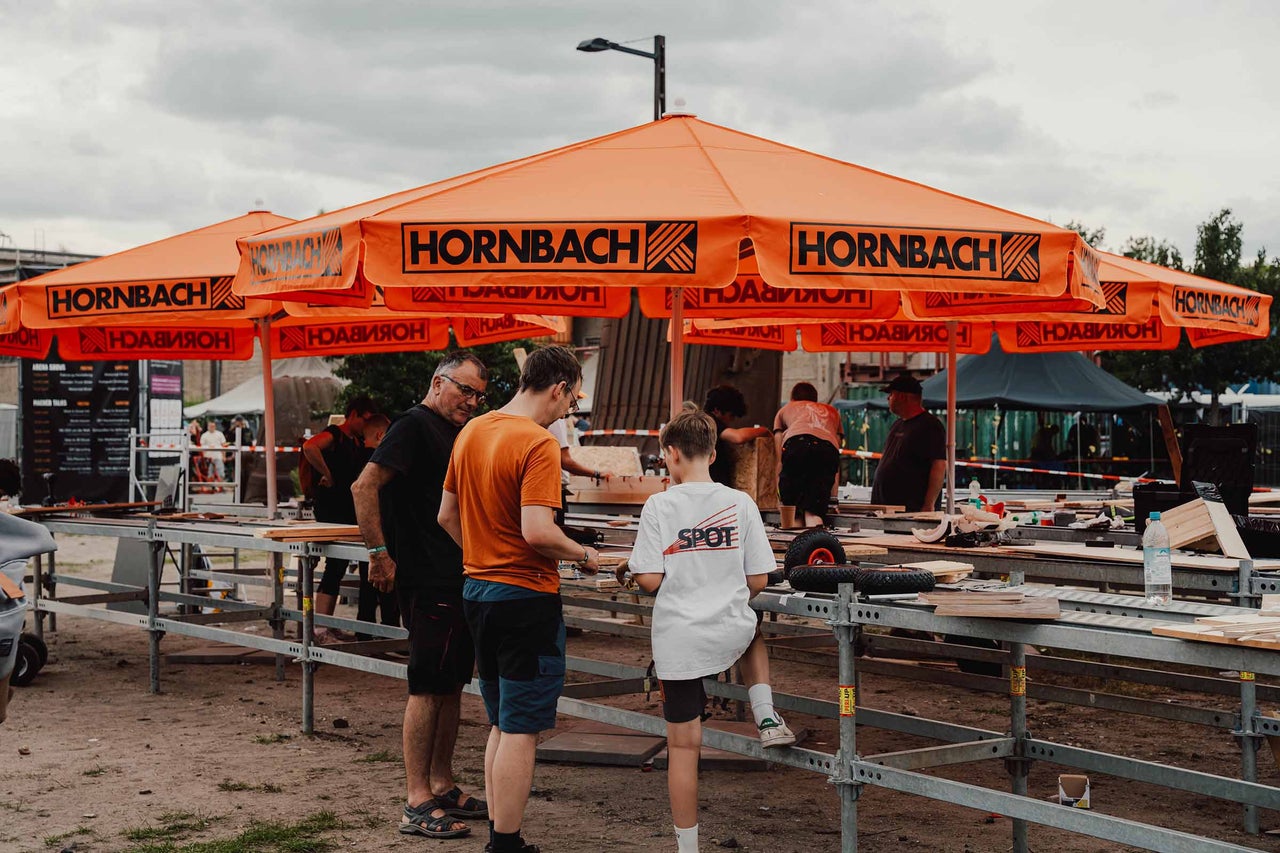 Szene im Freien mit Menschen, die an einem Tisch unter einem Sonnenschirm mit Hornbach Logo arbeiten.