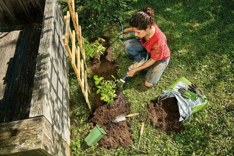 Frau gießt Pflanze im Garten mit Gartenschlauch, daneben Rankhilfe, Gartengeräte und Sack Blumenerde.