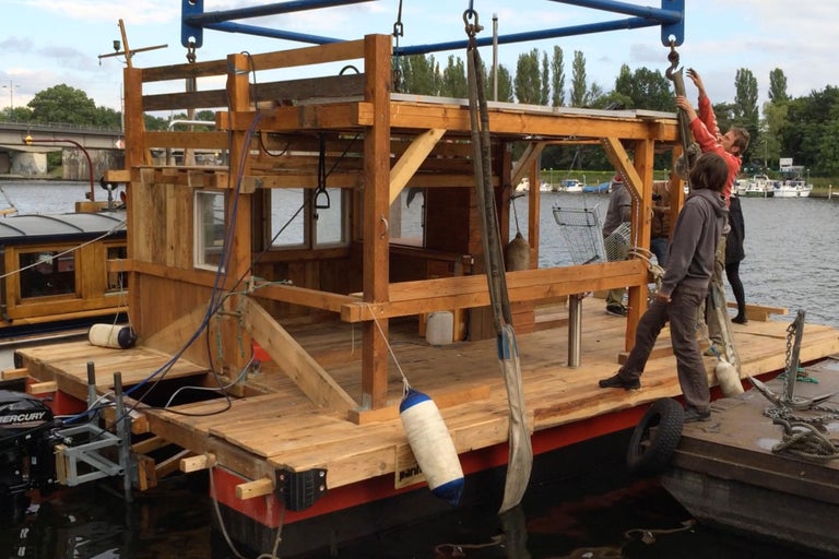 Ein schwimmendes Hausboot aus Holz mit Personen auf dem Deck.