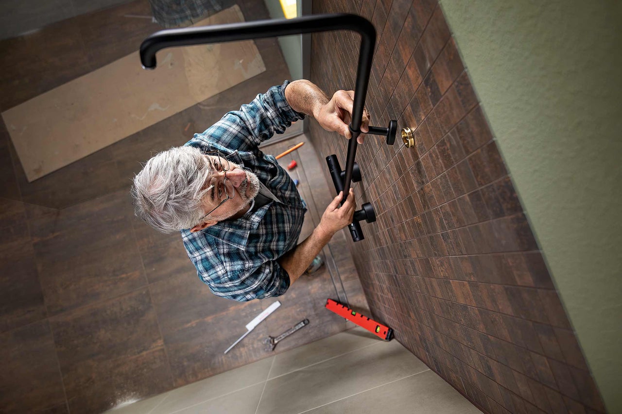 Ein Handwerker montiert eine Duschstange im Badezimmer an einer Wand mit braunen Fliesen.