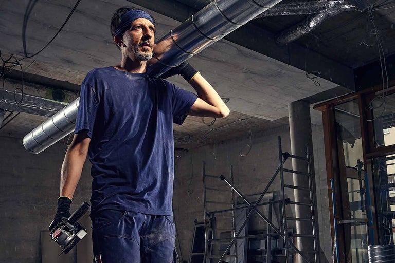 Ein Arbeiter trägt ein Lüftungsrohr auf der Schulter, im Hintergrund ein unfertiger Raum mit Gerüst.