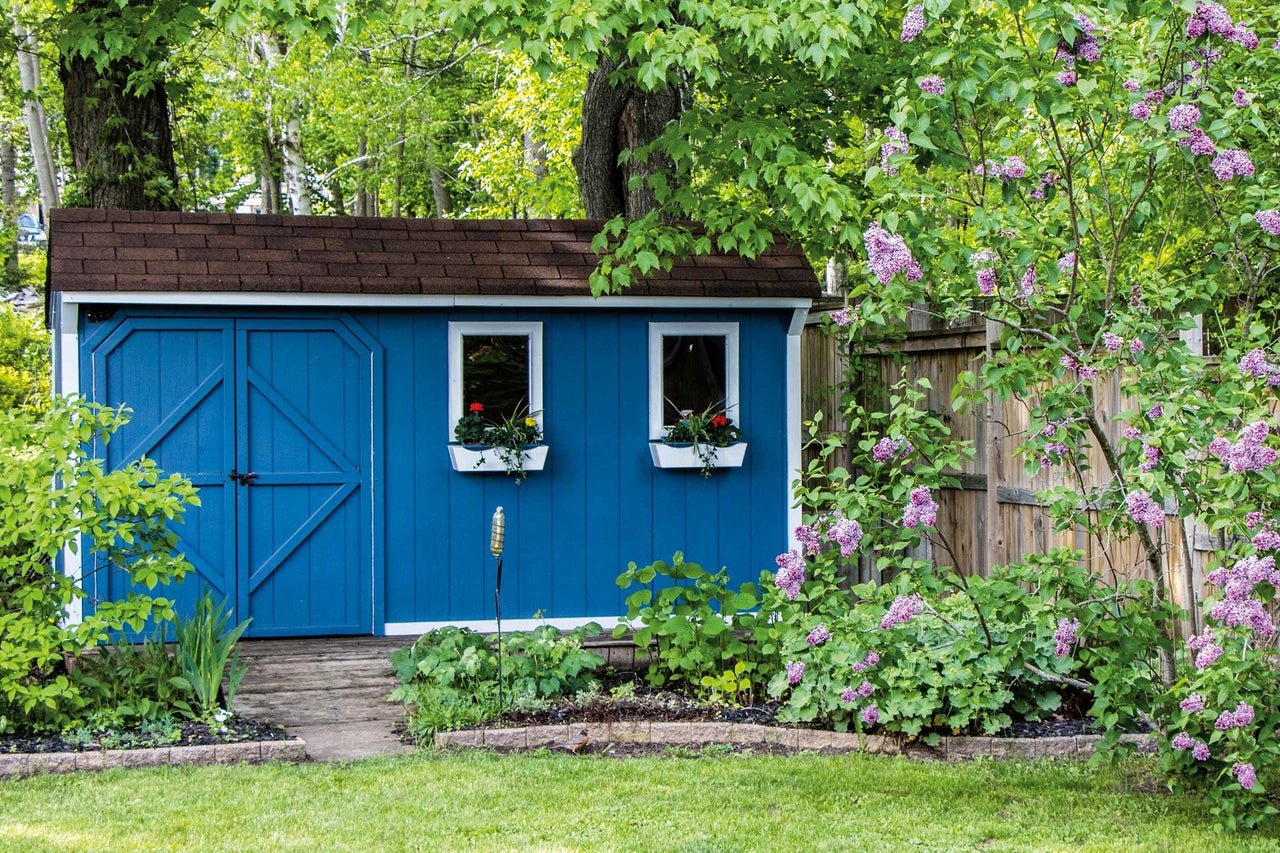 Gartenansicht mit blauem Geräteschuppen, Fenstern mit Blumenkästen und lila Blütensträuchern