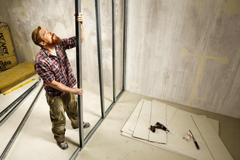 Ein Mann montiert Metallprofile für eine Trockenbauwand in einem Raum mit Gipskartonplatten, Isolierung und Werkzeugen.