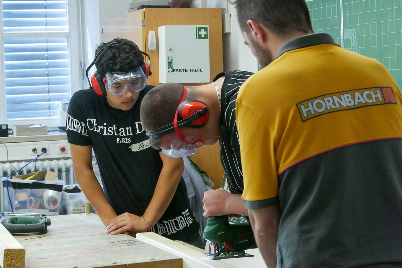 Jugendliche arbeiten mit einer Stichsäge an einem Holzstück, geschützt durch Schutzbrille und Ohrenschützer, Hornbach Logo sichtbar