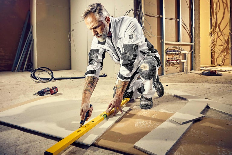 Ein Mann kniet in einem Raum im Rohbau und schneidet mit einem Cuttermesser eine Gipskartonplatte entlang einer Wasserwaage. Im Hintergrund sind Rohre, Kabel und eine Bohrmaschine zu sehen.