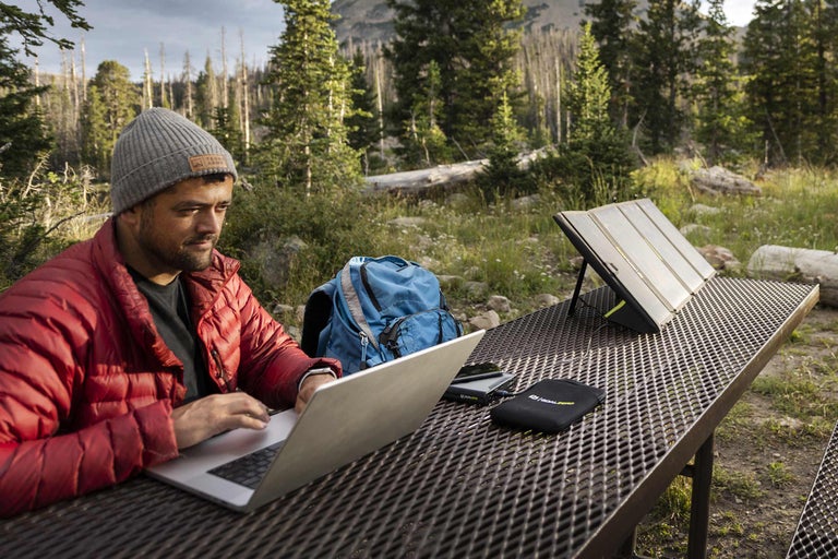 Mann arbeitet an einem Laptop mit einem tragbaren Solarmodul auf einem Tisch im Freien