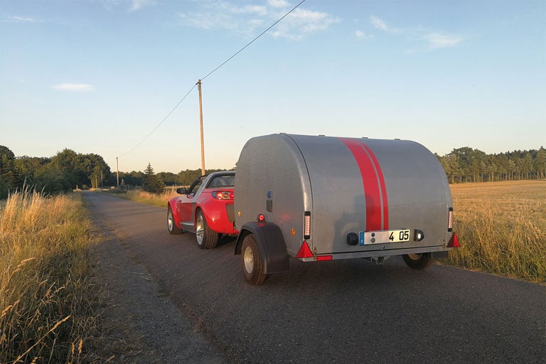 Ein rotes Auto zieht einen grauen Anhänger mit roten Streifen auf einer Straße.