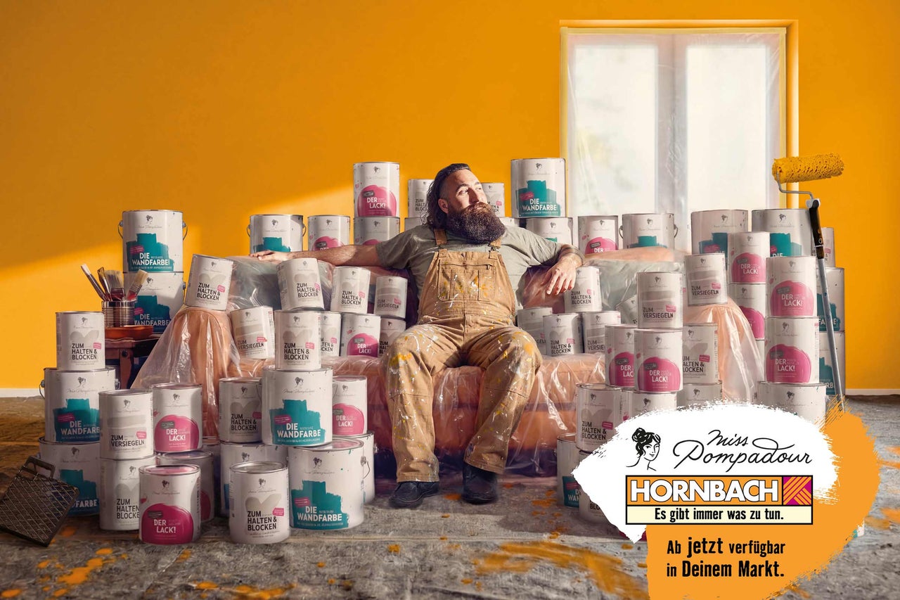 Mann in Latzhose sitzt auf Farbdosen in einem Raum mit gelber Wand und einem Farbroller, Miss Pompadour Hornbach Logo