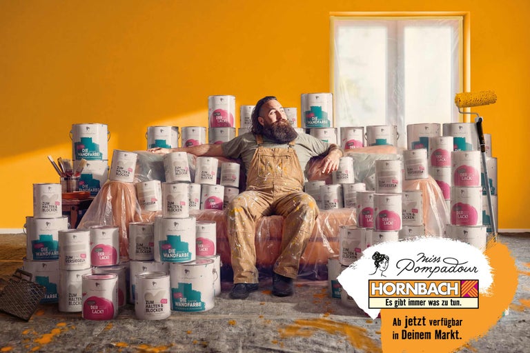 Mann in Latzhose sitzt auf Farbdosen in einem Raum mit gelber Wand und einem Farbroller, Miss Pompadour Hornbach Logo