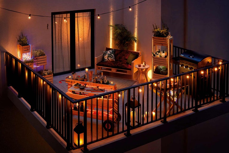 Atmosphärische Balkonszene mit Grill, Barwagen und Palettenmöbeln, dekoriert mit Lichterketten und Pflanzenkästen
