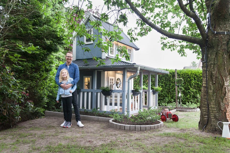 Ein Mann umarmt ein Mädchen vor einem blauen Spielhaus mit einem grauen Metalldach im Garten.