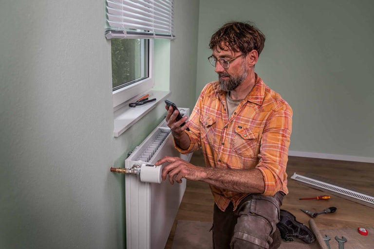 Mann installiert einen digitalen Heizkörperthermostat und verwendet ein Smartphone zur Steuerung.