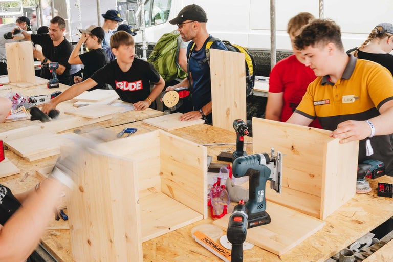Eine Gruppe von Jugendlichen baut gemeinsam Holzmöbel in einem Workshop mit Elektrowerkzeugen und Werkbänken unter Anleitung.