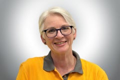 Lächelnde Hornbach Mitarbeiterin mit kurzen grauen Haaren und Brille in gelbem Poloshirt.