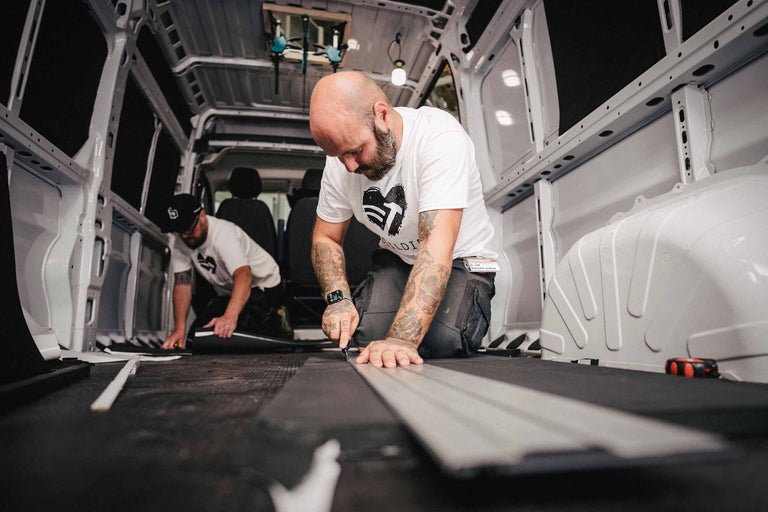 Zwei Handwerker schneiden Dämmplatten im Innenraum eines Lieferwagens zu.