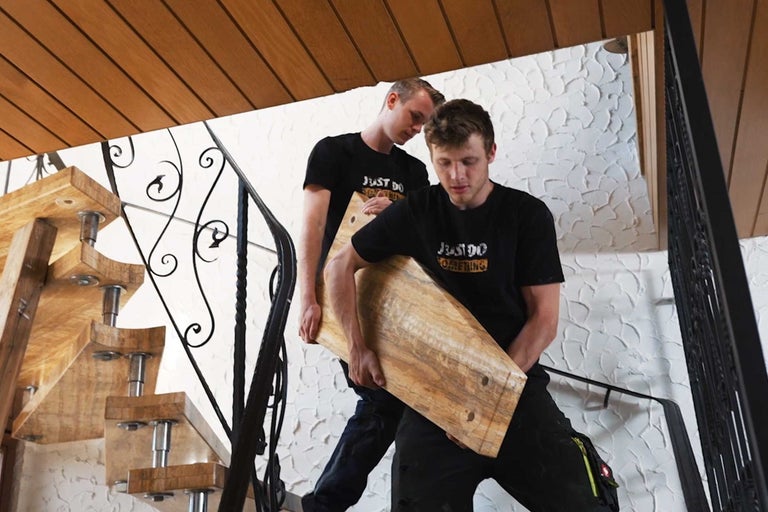 Zwei Personen tragen eine Holztreppenstufe auf einer Treppe mit Geländer.