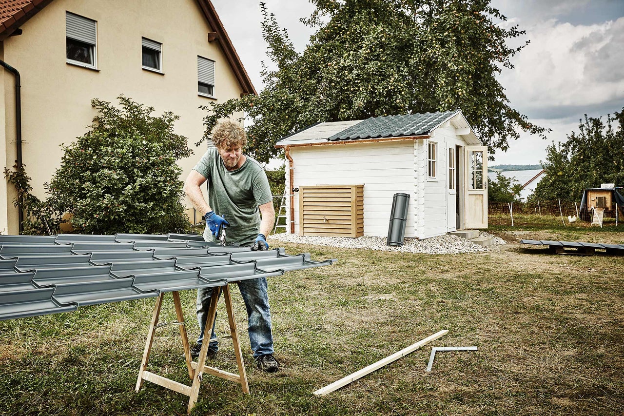 Mann schneidet Metall Dachplatten auf einer Holzkonstruktion im Garten, im Hintergrund ein Gartenhaus
