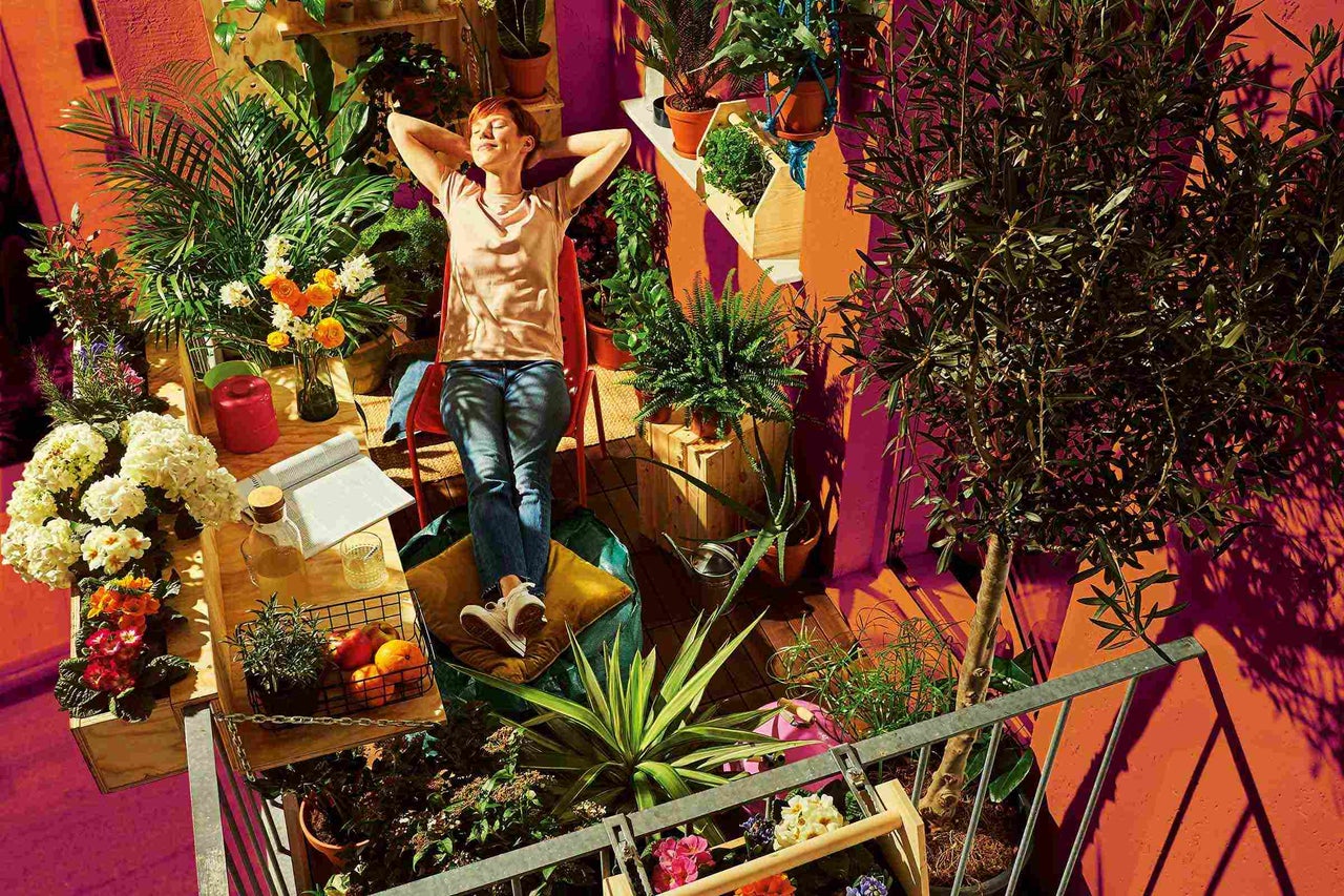 Frau entspannt auf dem Balkon, umgeben von Pflanzen, Blumen und Gartenmöbeln.