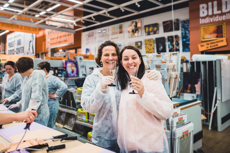 Zwei Kundinnen in Schutzanzügen und Handschuhen stoßen im Hornbach-Markt mit Sekt an.
