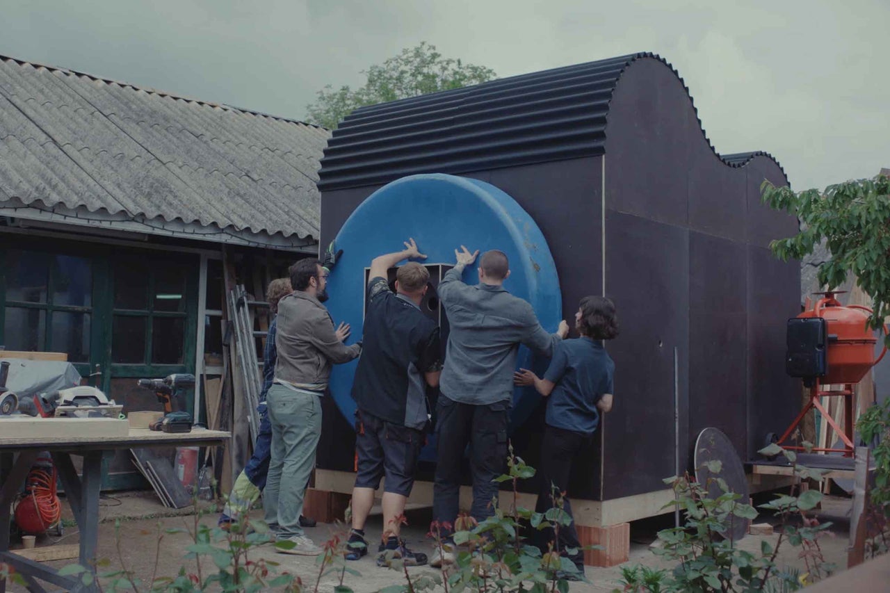 Eine Gruppe von Menschen montiert ein kreisförmiges Bauteil an die Außenwand eines modernen kleinen Holzhauses mit gewelltem Dach im Garten.