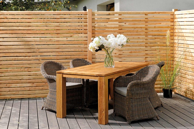 Szene mit Holztisch, Korbstühlen und Holzzaun auf einer Terrasse.