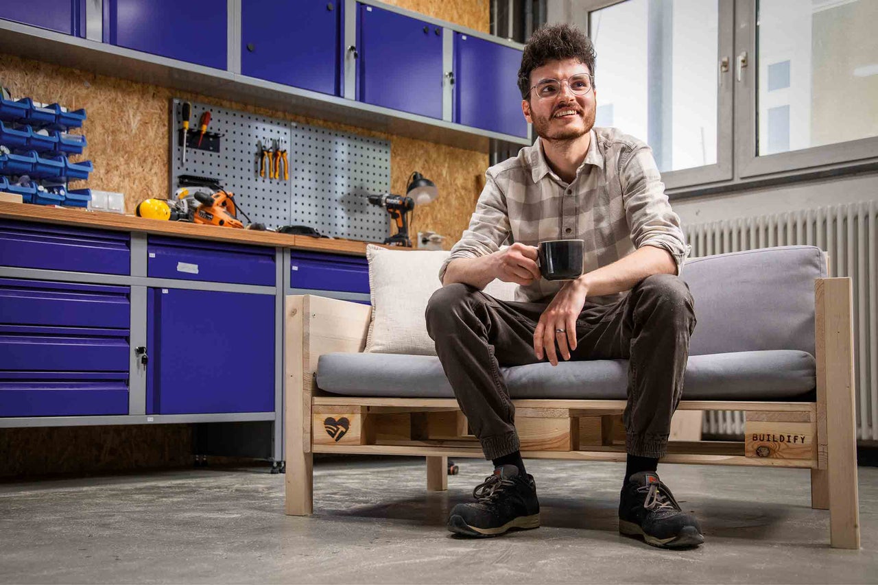 Mann sitzt mit einer Tasse auf einem Buildify Palettensofa aus Holz in einer Werkstatt mit Metallschränken und Werkzeugwand.