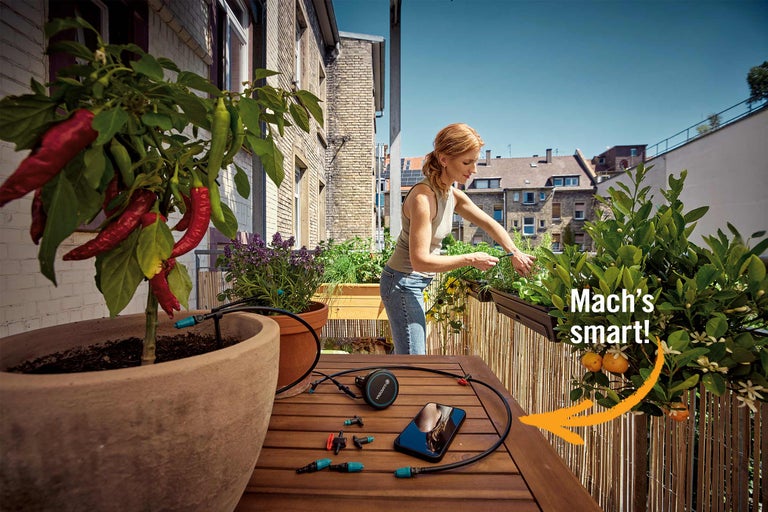 Eine Frau installiert ein smartes Bewässerungssystem auf einem sonnigen Balkon mit zahlreichen Topfpflanzen und Kräutern.