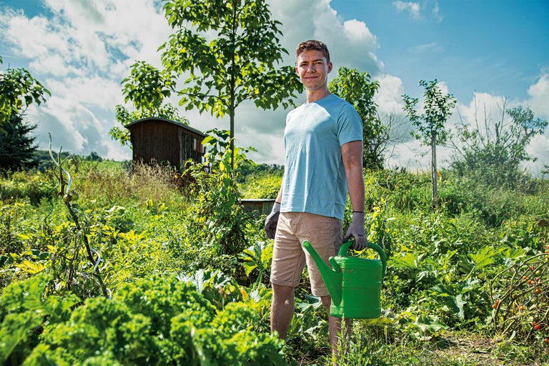 Ein Mann mit einer Gießkanne in einem Gemüsegarten mit Gartenhaus.