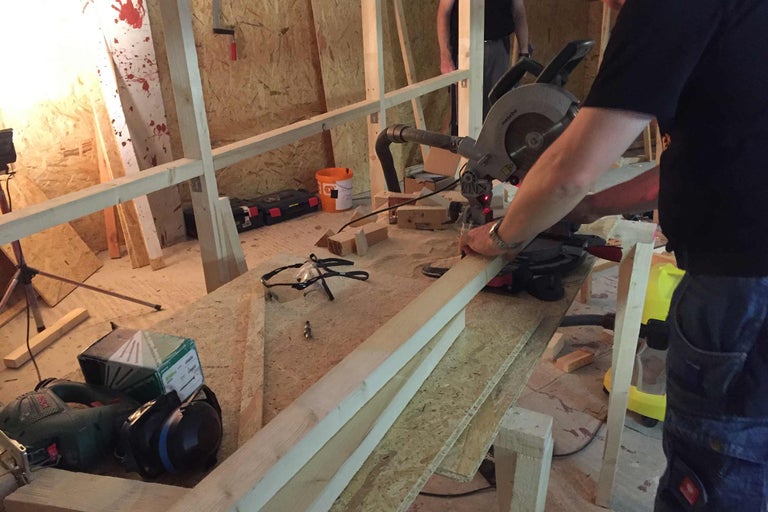 Person schneidet Holz mit einer Kappsäge in einer Werkstatt. Verschiedene Werkzeuge und Materialien sind im Raum verteilt.