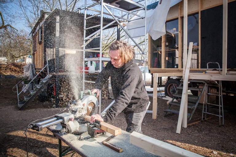 Eine Person arbeitet mit einer Kapp- und Gehrungssäge an einem Holzprojekt im Freien, mit einem Gerüst, einer Hütte und herumfliegenden Sägespänen.
