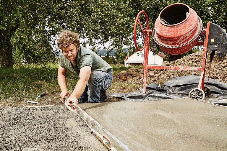 Mann glättet Beton mit Richtscheit neben Betonmischer