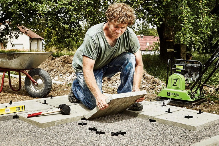 Mann verlegt Gehwegplatten mit einer Rüttelplatte, einer Schubkarre, einem Gummihammer und einer Wasserwaage im Garten.