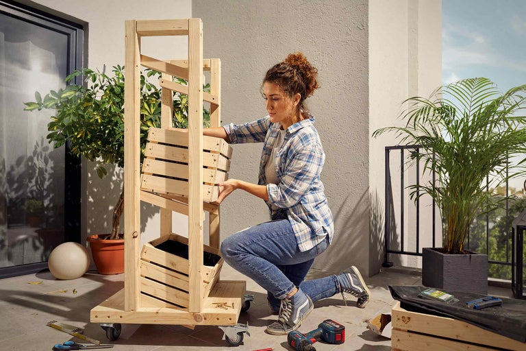 Frau montiert ein mobiles Hochbeet aus Holz auf einem Balkon mit Werkzeugen und Pflanzen.