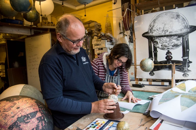 Zwei Personen arbeiten in einer Werkstatt an Globen und Karten