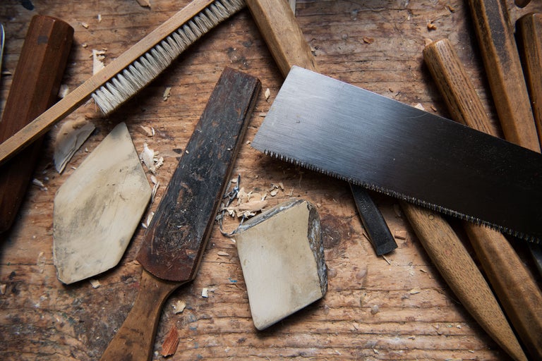 Verschiedene Werkzeuge wie Säge, Schleifstein, Pinsel und Stecheisen auf einer Holzoberfläche.