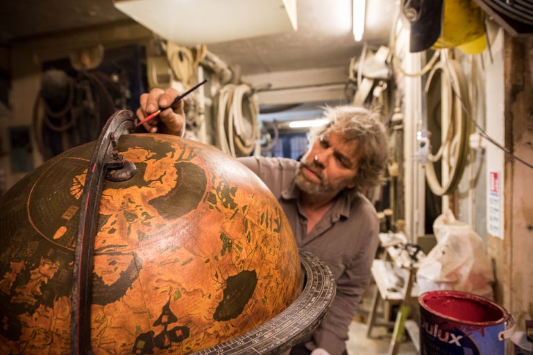 Ein Mann restauriert einen Globus mit einem Pinsel in einer Werkstatt.