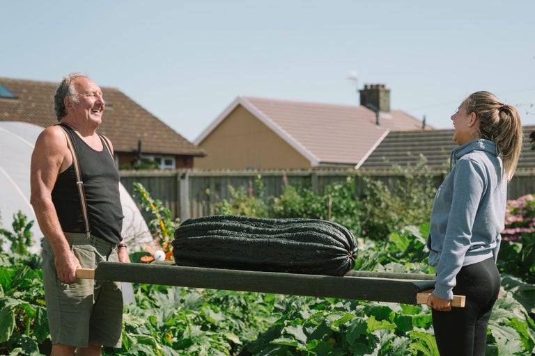 Zwei Personen tragen einen langen, dunkelgrünen Kürbis auf einer Trage durch einen Garten.