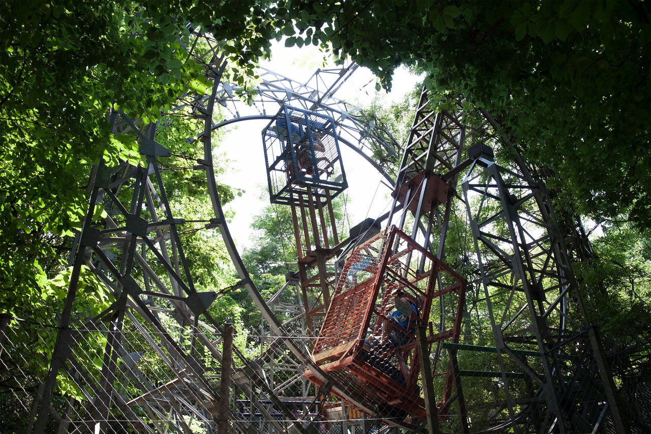 Blick von unten auf ein Riesenrad aus Stahl mit Fahrgästen in den Gondeln, umgeben von Bäumen.
