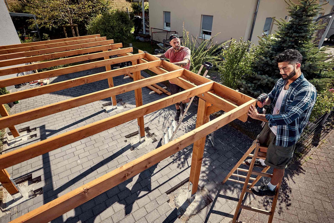 Zwei Handwerker montieren eine Holzkonstruktion für eine Terrassenüberdachung mit einem Akkubohrschrauber auf einer gepflasterten Terrasse im Garten.