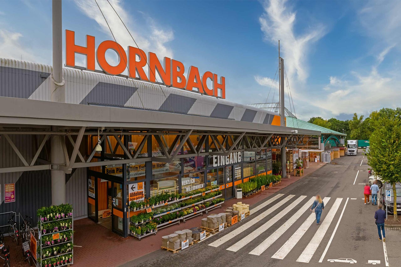  Aussenansicht eines Hornbach Baumarktes mit Eingang, Pflanzen und Parkplatz. 