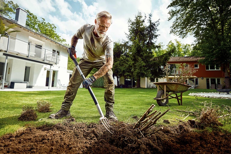 Ein Mann arbeitet mit einer Grabegabel im Garten, umgepflanzte Sträucher und eine Schubkarre im Hintergrund.