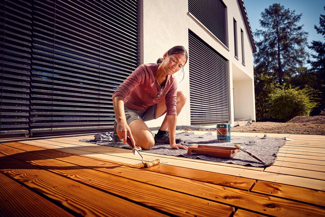 Eine Frau ölt eine Holzterrasse mit einer Farbrolle. Im Hintergrund ist ein modernes Haus zu sehen.