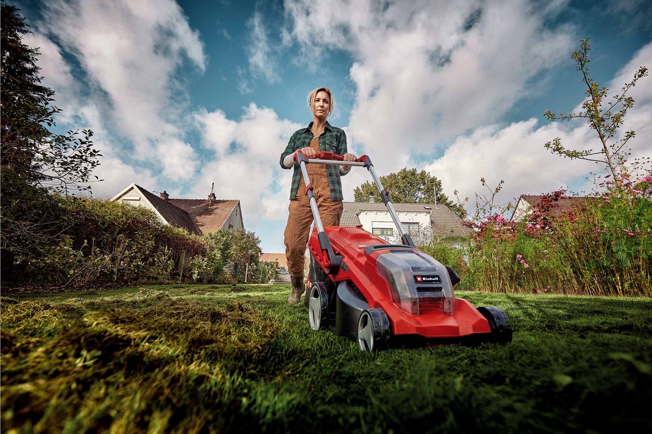 Eine Frau mäht mit einem roten Einhell Elektro-Rasenmäher einen grünen Rasen in einem Garten mit Häusern im Hintergrund.
