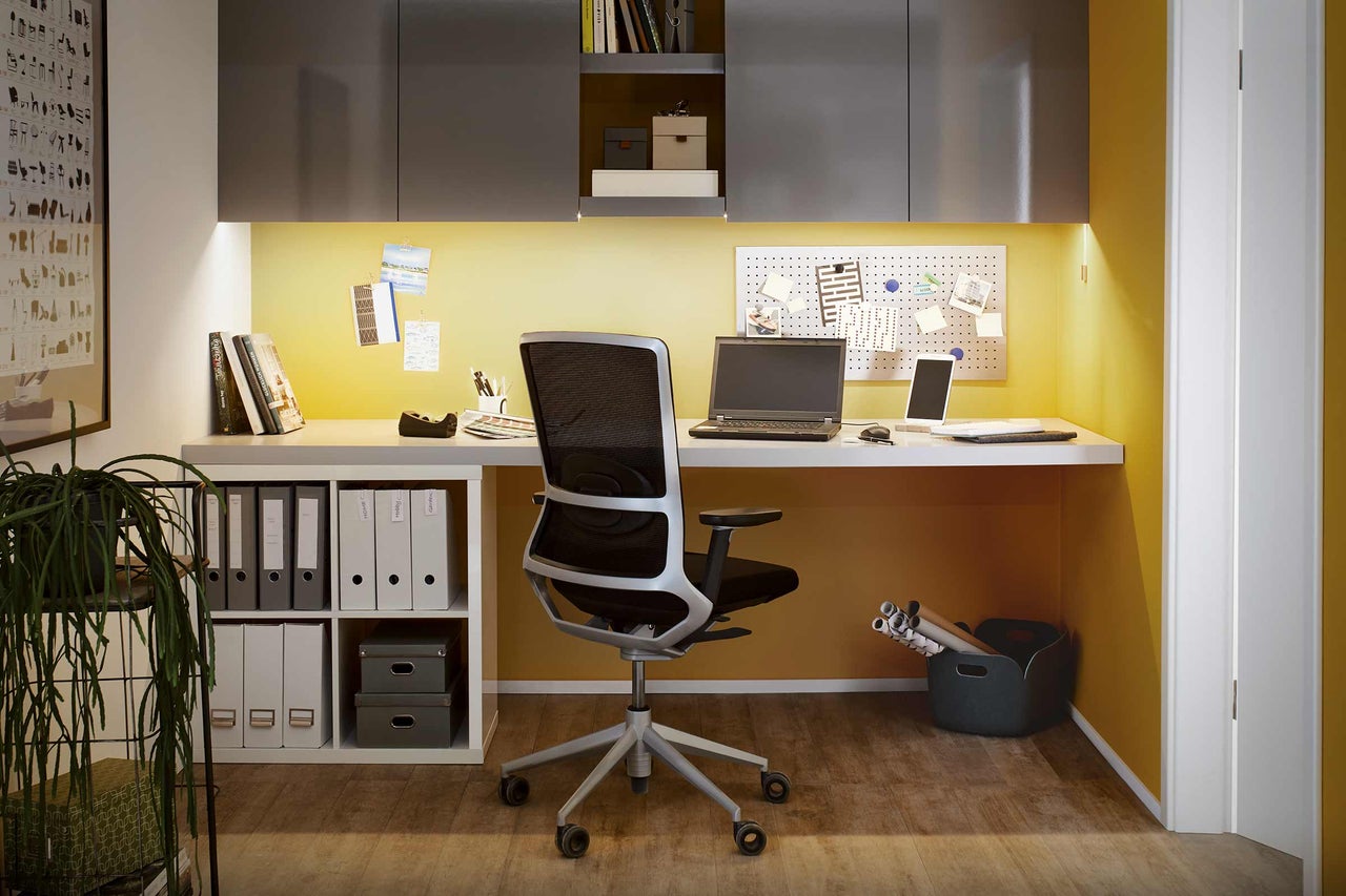 Büro mit Schreibtisch, Bürostuhl, Regalen und Arbeitsmaterialien in einem hellen Raum