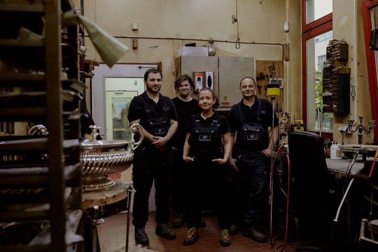 Vier Personen in Arbeitskleidung in einer Werkstatt mit Werkzeugen und Regalen.