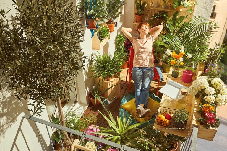 Frau entspannt auf einem begrünten Balkon mit Pflanzen in Töpfen und Blumenkästen.