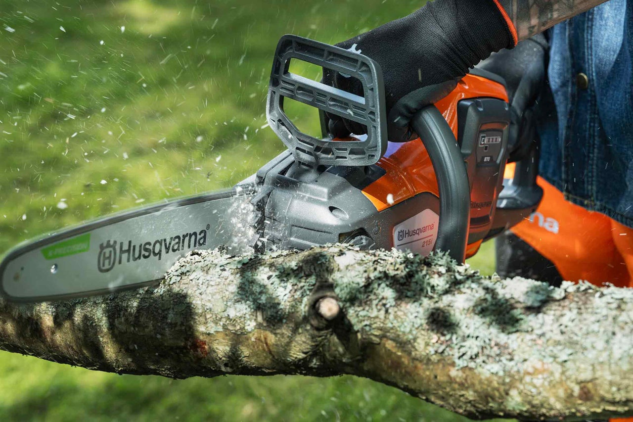 Husqvarna Akku-Kettensäge schneidet einen dicken Ast im Freien, fliegende Sägespäne, Husqvarna Logo.