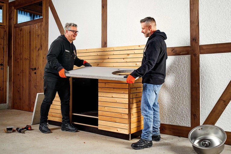 Zwei Handwerker montieren eine Gartenküche mit Holzverkleidung und Betonarbeitsplatte