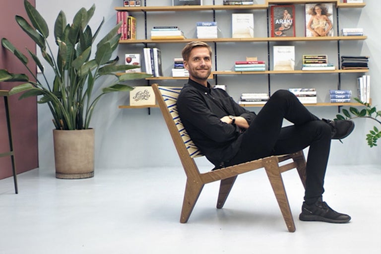 Mann sitzt auf einem Holzstuhl vor einem Bücherregal und einer Zimmerpflanze.